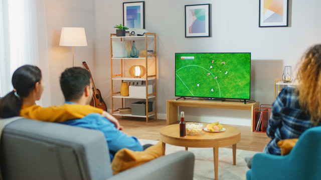 At Home Diverse Group Of Sports Fans Watches Important Soccer Match On TV, They Cheer For The Team. Cozy Room With Snacks And Drinks On The Table.