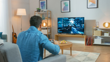 In the Living Room Man Sitting on a Couch Holds Controller Playing in a Console Video Game, 3D...
