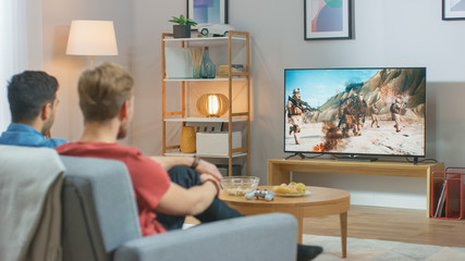In the Living Room: Two Friends Relaxing on a Couch Watching War Movie on a TV. Modern Military Warfare Action with Shooting and Explosions Shown on a Television.
