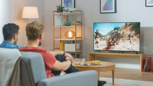 In The Living Room: Two Friends Relaxing On A Couch Watching War Movie On A TV. Modern Military Warfare Action With Shooting And Explosions Shown On A Television.