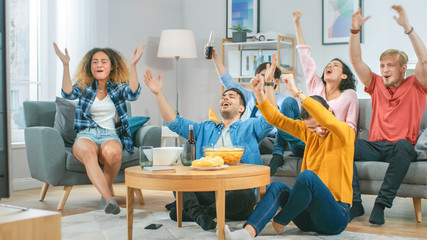 At Home Diverse Group of Sports Fans Sitting on Couch Watching Important Sports Game Match on TV, They Cheer for Team, Celebrate Victory after Team Scoring Winning Goal. Cozy Room with Snacks Drinks.