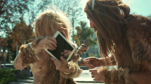 Prehistoric savage warriors discovering tablet computer device beating head with excitement in city outdoor. Ancient homo sapiens trying technology for first time.
