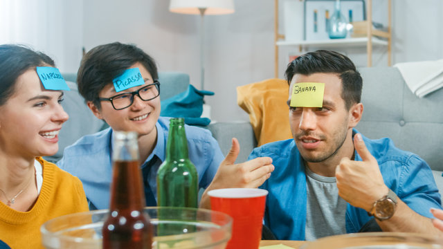 In the Living Room Diverse Group of Fun Loving Friends Playing "Who am I" game With Sticky Papers with Names on Written on them Attached to Foreheads. They Try to Guess What is Written on Sticky Notes