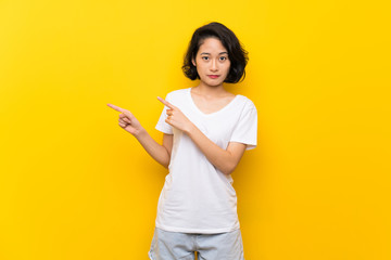 Asian young woman over isolated yellow wall frightened and pointing to the side