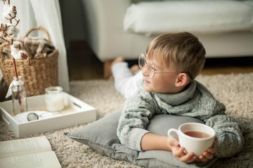 A beautiful little boy in a knitted sweater is reading by the window with a mug of hot tea. Cozy. Autumn.