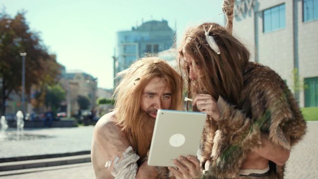 Prehistoric cave people of hunter-gatherers have fun browsing on modern tablet pc using internet first time staying in future city center. Human evolution.