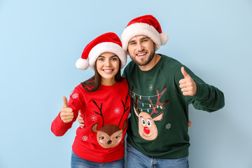 Happy couple in Christmas sweaters and Santa hats showing thumb-up on color background