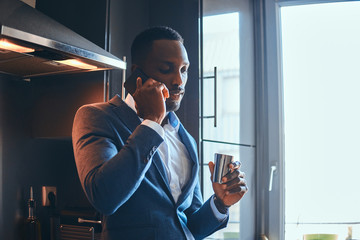 Elegant african man is enjoying his morning coffee while talking by mobile phone at his kitchen.