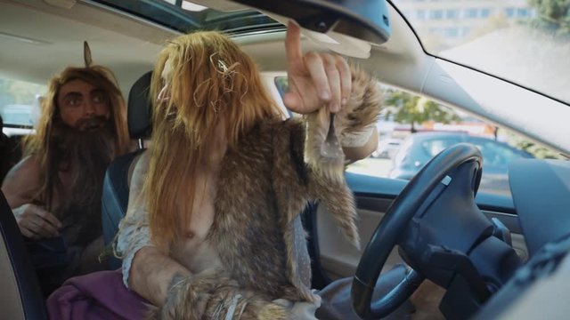 Two primitive neanderthals covered in animal fur looking in rearview mirror laughing while driving a modern car in city center. Homo sapiens.