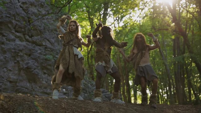 Funny neanderthals covered in animal fur dancing with excitement and joy on mountain rocks in jungle forest. Aboriginal lifestyle.