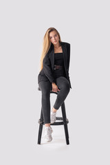 Fototapeta premium Full length portrait of beautiful young girl sitting on tall chair in black jeans and jacket over gray background