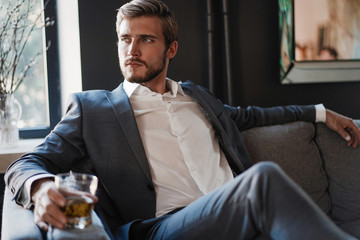 Handsome and successful businessman in stylish suit holding glass whiskey while sitting at office.