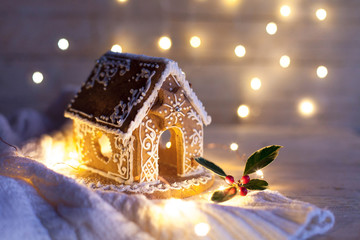 Christmas gingerbread house at night, cozy decorations on wooden and knitted background with glares. Cozy house in warm light with holly berries and cute white ornaments.