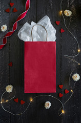 red gift bag on dark wooden table surrounded by holiday decor, flowers, twinkle lights, red hearts, red curled ribbon, blank gift wrap, copyspace, holiday flat lay