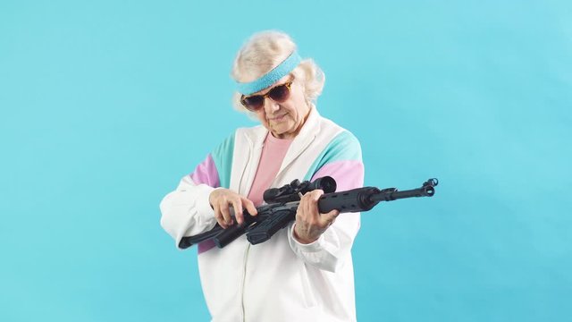 Old woman in sportswear and black sunglasses is holding a rifle isolated on blue.