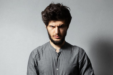 Obraz premium Portrait of bearded young guy with disheveled hair, looking angry at camera, on background of grey.