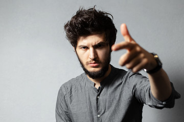 Portrait of young bearded and serious guy with disheveled hair, pointing with finger towards the camera, on grey background.