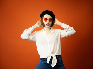 Fototapeta na wymiar Young woman with scared face holding her head Basque Beret, red glasses and white blouse, short hair isolated on orange studio. Beautiful female in posing. Advertising Person. Lifestyle concept