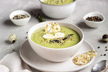 Detox broccoli green cream soup with seed mix, quail egg and crackers in bowl on light grey stone background. Healthy food concept.