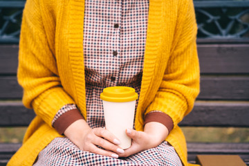 A woman is sitting on a park bench and holding a bamboo cup with hot coffee in her hands. Fashionable autumn clothes in orange mustard tones