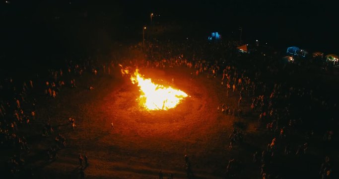A large crowd of people celebrate near a giant bonfire. Burning hot sparks rise from large fire in the night sky. Belarusian Kupalle. Russian ethnic holiday Ivan Kupala in village. Aerial drone shot.