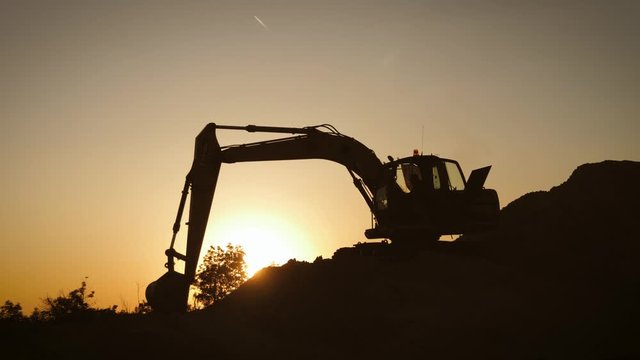 Silhouette of an excavator that loads sand into a truck at sunset. Concept construction and heavy industry, machine will be used in heavy industry business. Slow motion footage.
