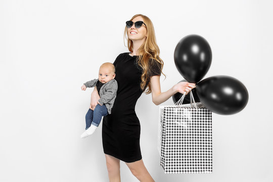 Happy elegant young mom, girl in black dress happily holding bags with black balloons, and little baby on white background. Baby shopping
