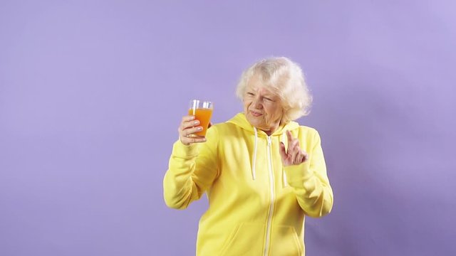 Joyful old woman in a yellow sports sweatshirt holds a glass of fresh carrot juice in her hand.