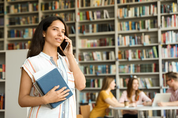 Young woman with books talking on phone in library. Space for text