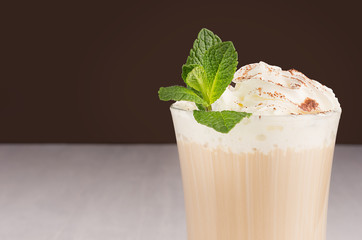 Latte coffee closeup, top section of glass with whipped cream, green fresh mint and cocoa powder, details on dark brown background, copy space.
