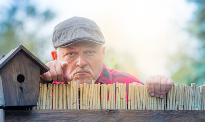 An elderly man in a hat looks angry and watches the neighborhood over a garden fence. Concept:...