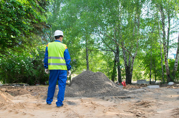 a worker in a blue uniform and a yellow vest, stands with his back in front of a pile of soil to repair the road