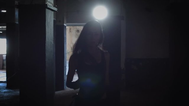 Asian girl with long dark hair dancing in abandoned warehouse in a black swimsuit, latex shorts, latex gloves against backlight.  Focus pulling, moving camera