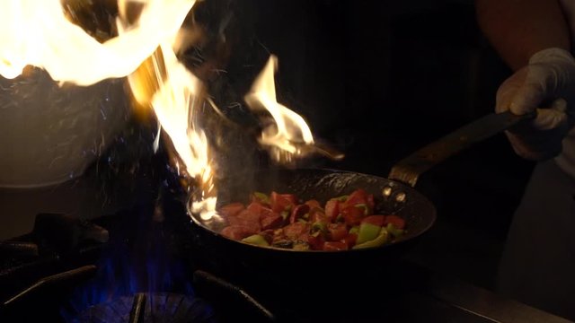 Chef frying vegetables on fire throwing them in a frying pan. Slow motion view of frying pans with vegetables on big fire flame at street cafe in Turkey. Cook frying vegetable in the kitchen