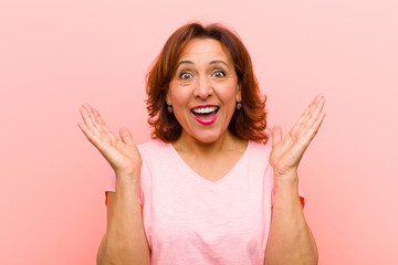 middle age woman looking happy and excited, shocked with an unexpected surprise with both hands open next to face against pink wall
