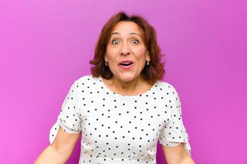 middle age woman looking very shocked or surprised, staring with open mouth saying wow against purple wall
