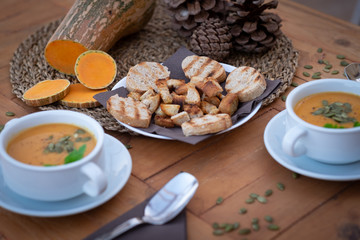 Close-up of two bowls of pumpkin cream soup. In the middle of the wooden table bread croutons.