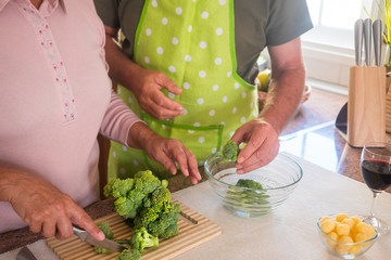 Pair of old human hands cleaning and cutting broccoli. Two people working together. Healthy and vegetarian food. Wooden cutting board