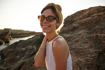 Young attractive dark haired woman in vintage sunglasses and headband posing over promenade on early summer morning, keeping hand on her neck and smiling cheerfully to camera