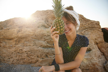 Outdoor sunny photo of charming young brunette lady in summer dress and headband sitting over yellow stone, holding fresh pineapple in hand and smiling to camera happily - Powered by Adobe