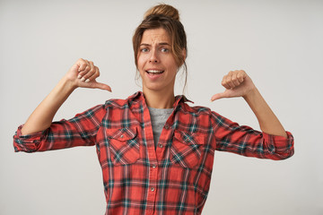 Fototapeta premium Indoor shot of attractive young self-confident woman with dark hair posing over white background in casual clothes, showing to herself with thumbs