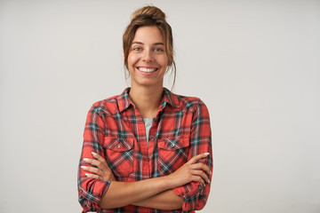 Studio portrait of young pretty dark haired female with bun hairstyle wearing casual clothes, posing over white background, looking happily to camera with crossed arms on chest - Powered by Adobe