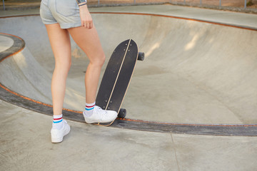 Cropped outdoor photo of slim female in jeans shorts posing over skateboard on summer warm weather,...