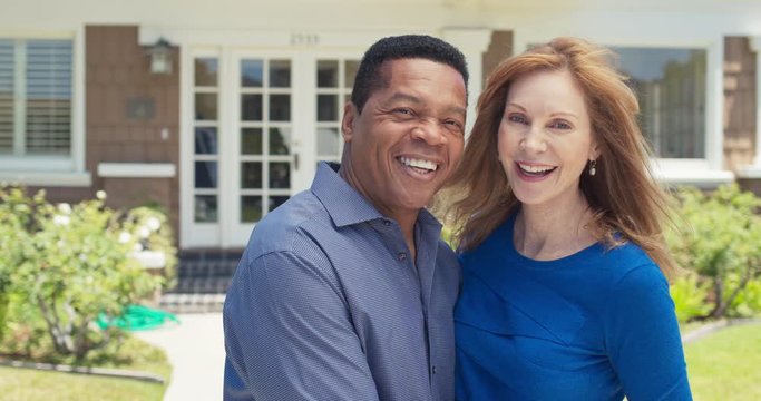 Portrait of smiling older African American and Caucasian couple standing in front of their home. Happy senior husband and wife homeowners looking at camera outside house. Slow Motion 4k Handheld