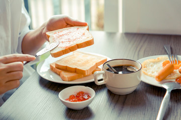 Breakfast - Hand spreading strawberry jam on toast with coffee on table, sausage, and fried egg