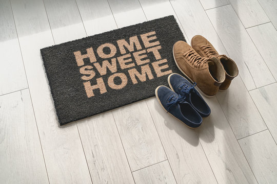 Home sweet home doormat at condo door entrance with couples pairs of shoes moving in together. women's sneakers and man's boots on floor, new apartment.