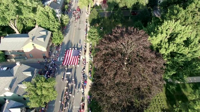 Aerial of crowds lining streets of small town parade, US flags fill grassy lawn, flag carried by Boy Scouts of America