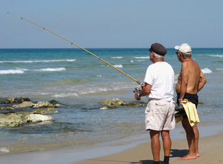 Two tanned elderly men fishing with a fishing rod in the sea from the shore