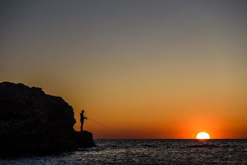 Sea, sunset, and the silhouette of a fisherman-the silhouette of a man with a fishing rod on the background of the setting sun over the sea.