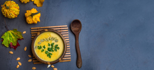wooden bowl of traditional pumpkin soup on gray table, decorated with seeds, parsley, colorful foliage, decorative pumpkin
